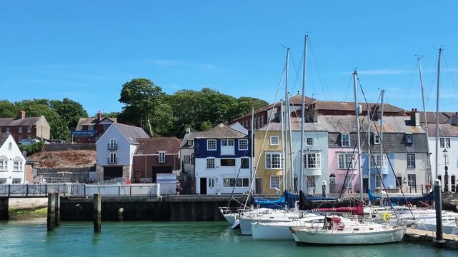 Weymouth Dorset England UK.  02.07.2026. Video. A picturesque coastal town scene featuring vibrant houses lining a harbour filled with docked sailboats under a clear blue sky