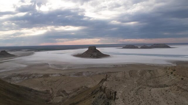 A spectacular cinematic drone shot flying over a steep rocky cliff edge, revealing a massive isolated rock monolith standing in the shallow, reflective waters of Karynzharyk  kazakhstan