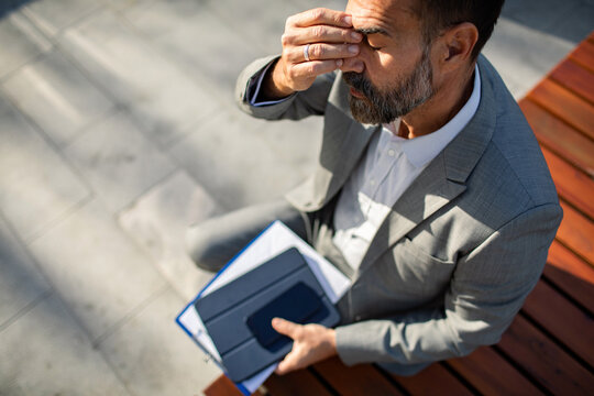 Stressed businessman rubbing eyes with headache on outdoor city bench