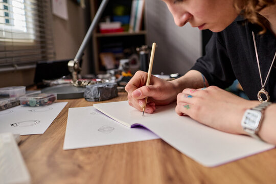 Close up of young adult Caucasian woman sketching jewelry design in notebook at workbench, developing handmade product concept. Ideal for artisan branding, creative business marketing