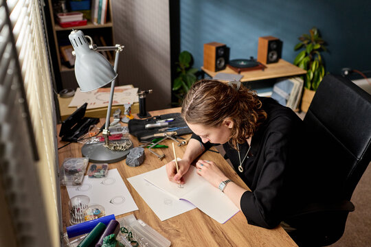 High angle view of young adult Caucasian woman sketching jewelry design at workbench, developing handmade product concept. Ideal for artisan business, creative entrepreneurship marketing