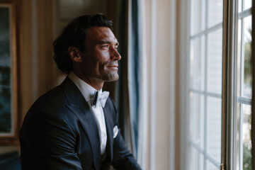 Fototapeta premium A groom dressed in a classic tuxedo gazes out the window, capturing a moment of reflection and sophistication as he prepares for the joyful wedding celebration ahead.