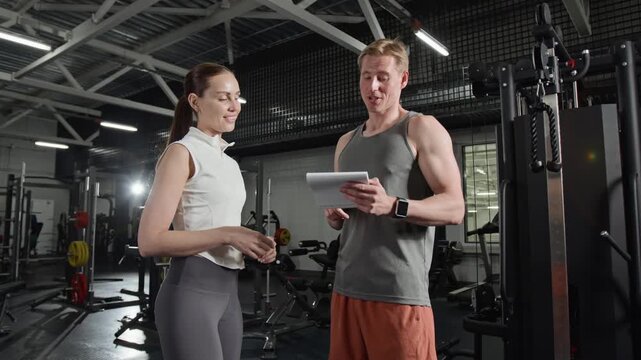 Young male fitness trainer holding notebook and explaining workout plan to female client in gym. Clip for promoting personal training services