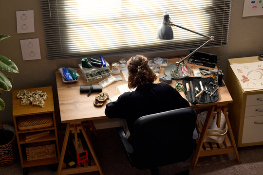 High angle view of young adult woman crafting handmade jewelry at workbench, arranging tools and materials for small business production. Ideal for artisan branding and ecommerce marketing