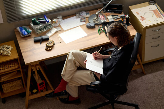 High angle view of young adult woman sketching jewelry design at workbench, developing handmade product concept for creative business. Ideal for artisan branding and ecommerce marketing