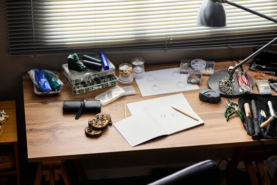 High angle view of jewelry maker workspace displaying sketchbook, design drafts, hand tools, metal findings, supporting artisan production and small business branding or ecommerce content