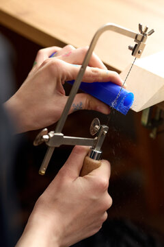 Vertical shot of young adult craftsperson hands sawing blue wax tube at jeweler bench, shaping prototype for handmade product development. Ideal for artisan training or small business marketing