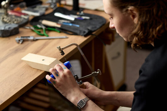 Young adult woman shaping jewelry piece with coping saw at workbench, practicing handcraft production and precision skills. Useful for artisan business, maker education, creative workshop marketing