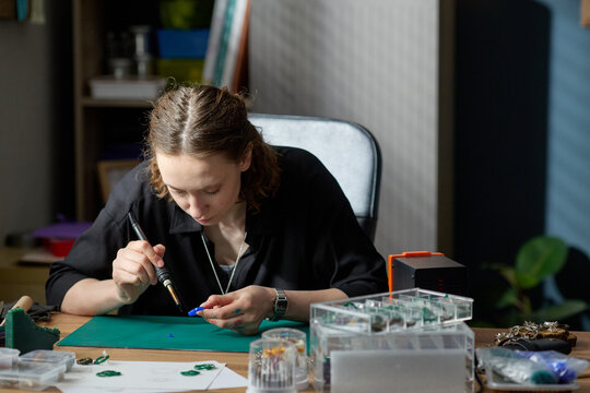 Young adult Caucasian woman soldering small component at workbench, assembling handmade product for small business production. Useful for maker economy, craft startup, ecommerce content