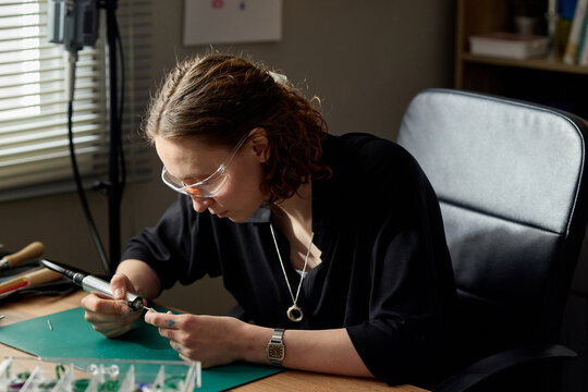 Young adult Caucasian woman crafting jewelry at workbench, using hand tool with precision and focus. Ideal for artisan business, handmade product marketing, creative entrepreneurship