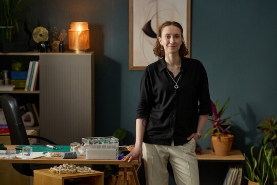 Portrait of young adult Caucasian woman standing beside desk, looking at camera in creative workspace, presenting small product design process. For branding, startup, maker business marketing