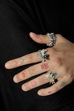 Vertical shot of young adult woman hand displaying statement rings and playful temporary tattoos against black fabric. Supporting jewelry branding, accessory retail, fashion campaign visuals