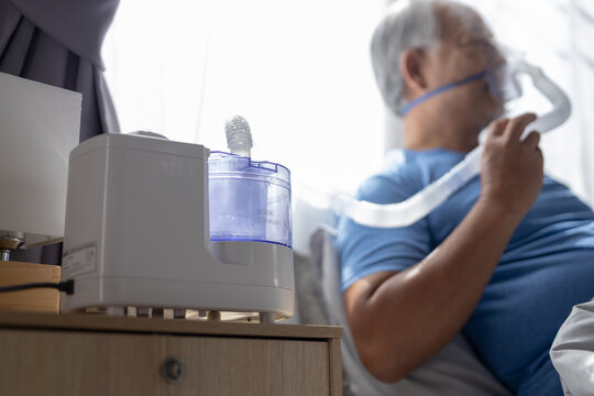 Senior man using cpap machine wearing oxygen mask sleeping comfortably in bed with peaceful expression resting calmly breathing easy during daytime at home clinic bedroom interior feeling relaxed