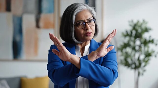 Mature woman with short gray hair and glasses making a crossing gesture with her arms to signal no or stop, expressing rejection or refusal through body language in studio.