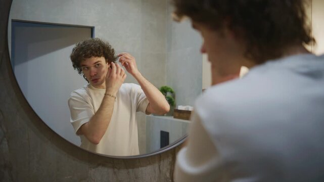 Young man styles his curly hair while looking in a round mirror in a bathroom during the evening hours