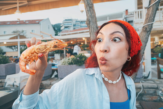 Woman with red hair, wide eyed and mouth open, holding a big shrimp with a surprised expression in a restaurant setting