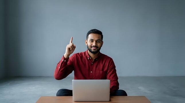 Young man with dark hair and beard, South Asian ethnicity, wearing burgundy button-up shirt, sitting at desk with laptop, pointing upward with index finger in eureka gesture, studio setting.