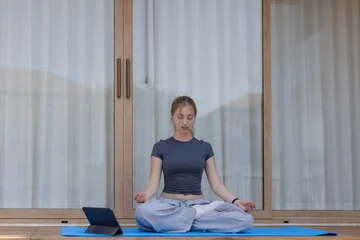 Fotobehang Lotusbloem Young peaceful woman meditate on blue yoga mat using tablet for health outdoor at home. Beautiful calm girl sitting in lotus pose outside glass door enjoying relaxing wellness morning routine  © Ratirat