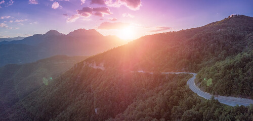 Obraz premium Winding mountain road traversing a lush green forest with dramatic sun flare and mountain peaks during a vibrant sunset