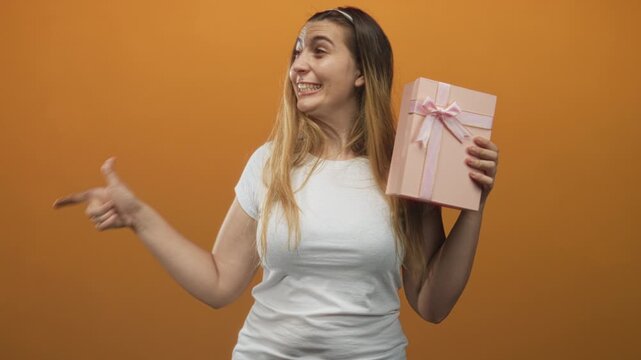 Woman in white tshirt holds pink gift box with bow and points right index finger to side in studio; joyful surprise celebration.
