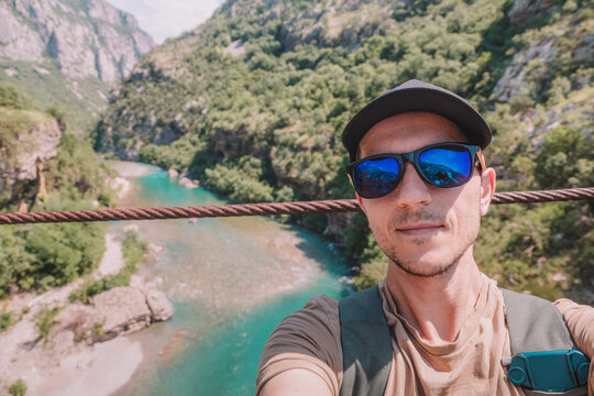 Young man standing on a bridge, enjoying adventure travel and beautiful natural canyon landscapes. Reflecting mountains in sunglasses