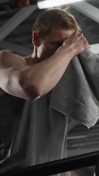 Vertical low angle video of young Caucasian man running on treadmill, wiping sweat with towel, and drinking water. Clip for gym promotions and healthy lifestyle marketing campaigns