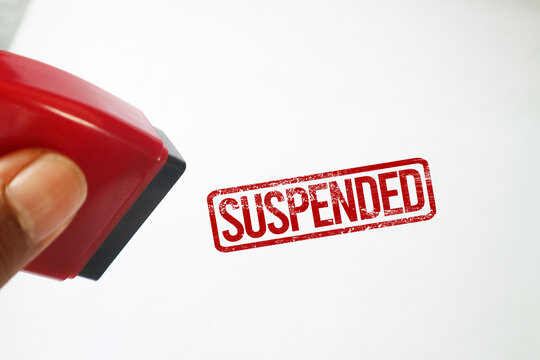 A hand holding a red rubber stamp after making a SUSPENDED imprint on a white surface.