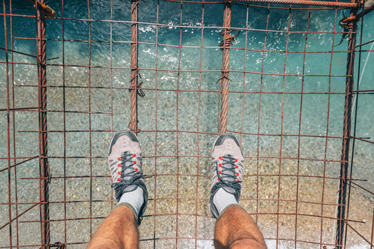 Adventurer is standing on a shaky old suspension bridge, looking down at the clear blue water rushing below, feeling height and thrill