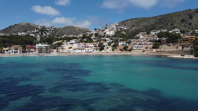 Cinematic drone shot flying away from El Portet beach in Moraira, Spain, revealing luxury villas nestled on the hills and white yachts anchored in the crystal-clear turquoise bay.