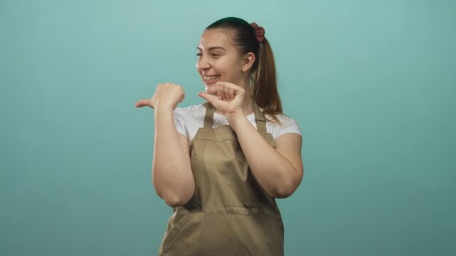 Woman in apron points thumb to chest while smiling and looking aside in studio, clear self referral gesture; confidence.