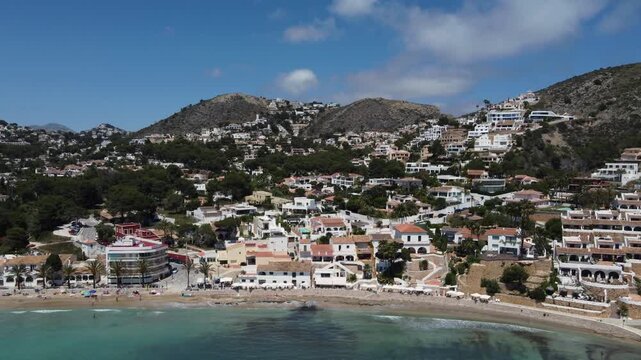 Aerial drone shot ascending and flying away from the scenic shores of El Portet beach in Moraira, Spain, revealing the beautiful Mediterranean coastline.
