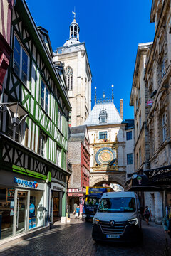 Urban street scene in Rouen, France, showing the Gros Horloge Renaissance clock tower rising above a busy street with delivery vans, green and red half-timbered buildings,.