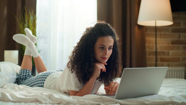 Serious pensive concentrated Caucasian woman using laptop lying on bed browsing computer female girl thinking thoughtful look in computer browsing device distant education online shopping in bedroom