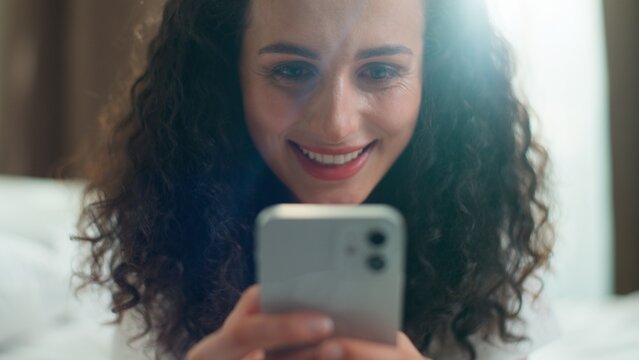 Positive Caucasian woman relax in bedroom home addicted overuse smartphone gadget internet social media website at morning waking up at bed sunny apartment smiling girl happy female using mobile phone
