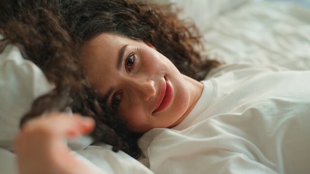 Close up Caucasian pretty woman lying in cozy bed looking at camera happy smiling pampering flirting at bedroom beautiful female girl with healthy skin resting calm peaceful at home hotel at morning
