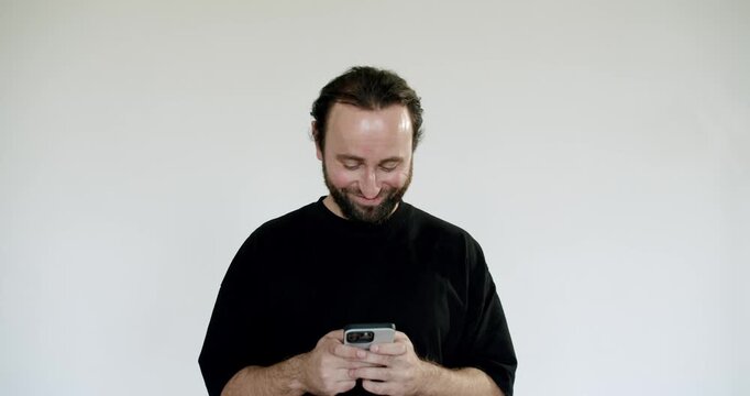 A man with a beard and long hair tied back is standing against a plain off-white wall. He is looking at his phone and smiling. He is wearing a black t-shirt.