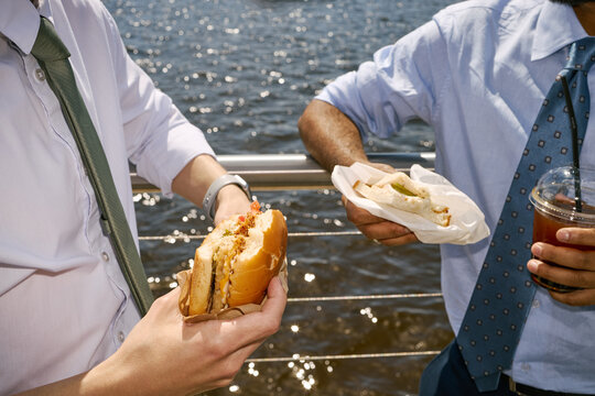 Indian and Caucasian businessmen eating lunch outdoors during work break, holding sandwiches and iced drink. Supporting business lifestyle, team bonding, workplace wellness marketing