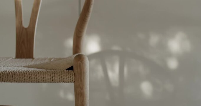 Close up of a light wooden chair with a woven seat. Sunlight streams in from a window, casting elegant shadows from tree branches on a simple wall behind. Minimalist furniture design.