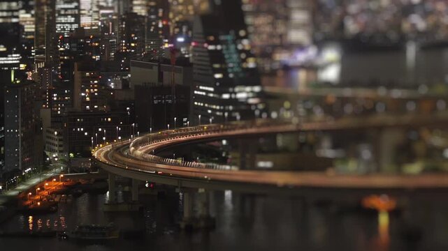 Night timelapse of an elevated highway curve with traffic light trails above a canal and dense city buildings in Tokyo, Japan. Tilt shift miniature effect and urban bokeh lights.