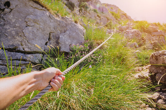 Hiker's hand firmly gripping a rope for support while ascending a steep, rocky trail, moving upwards in nature