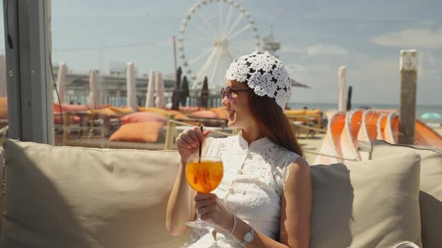 A woman in a white lace dress and sun hat holds an orange cocktail at a beach club.