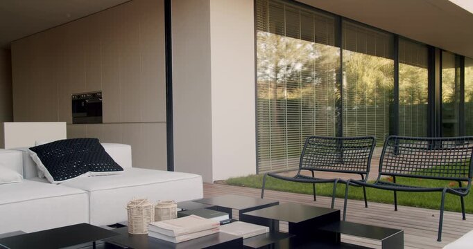 Modern home backyard patio area featuring white sofa, black wicker chairs, black square tables, wood deck, grass lawn, and glass sliding doors with blinds and trees reflecting in the glass.