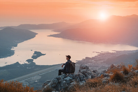 Man with backpack sitting on rocks, watching sunset over the calm waters of Bay of Kotor fjord landscape from a high mountain in Montenegro