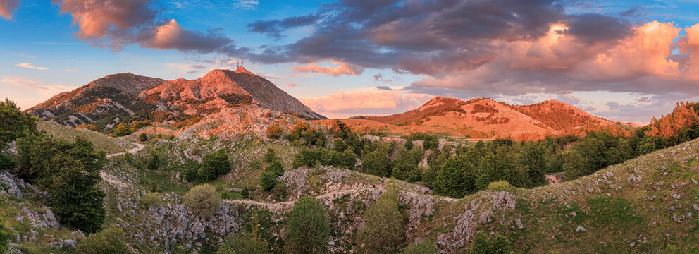 Rugged Lovcen mountain peaks and rolling hills illuminated by warm sunset light, with winding paths in Montenegro