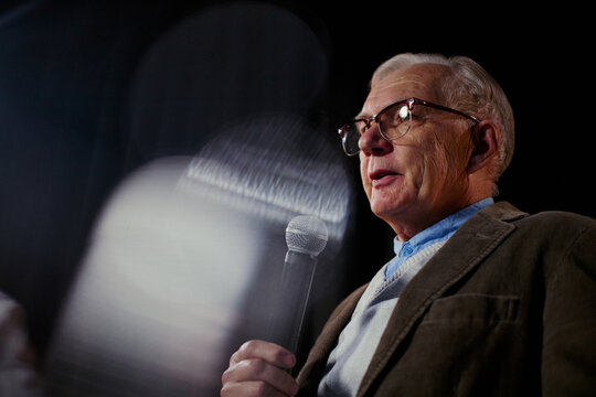 Senior Caucasian man speaking into microphone on stage, presenting leadership message during public event. Useful for corporate communication, keynote marketing, expert branding