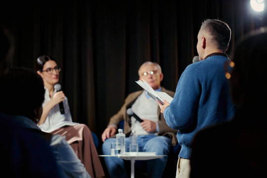 Young adult man speaking into microphone and reading notes during panel discussion, engaging speakers on stage. Useful for conference marketing, public speaking, leadership content