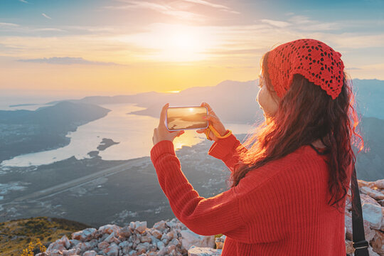 Young woman standing on a mountain peak, enjoying a beautiful sunset over the Bay of Kotor while recording the view with a mobile phone