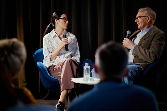 Medium shot of Caucasian businesswoman and senior man speaking on stage during panel discussion, sharing expertise and leadership insights. Ideal for conference marketing and corporate communication