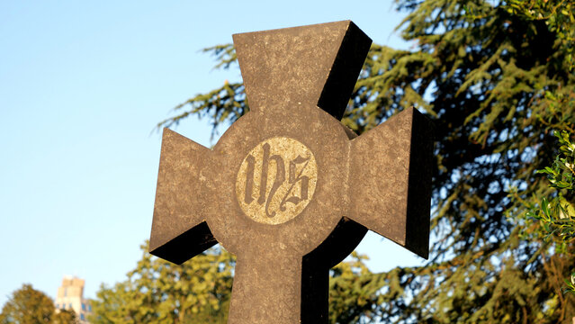 Stone cross monument with IHS monogram, historic christian religious symbol in cemetery, weathered grave headstone monument with green trees and clear sky