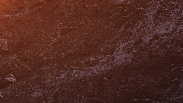Waves roll onto the beach as the sun sets. The water pushes against the sand, forming shapes and patterns. The sky turns orange, adding color to the scene and reflecting on the surface.